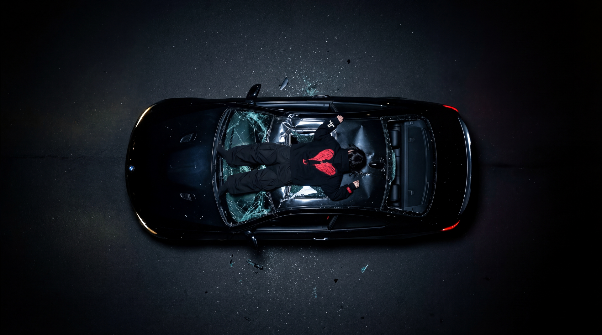 Overhead view of model lying on a smashed black car windshield wearing a broken heart hoodie